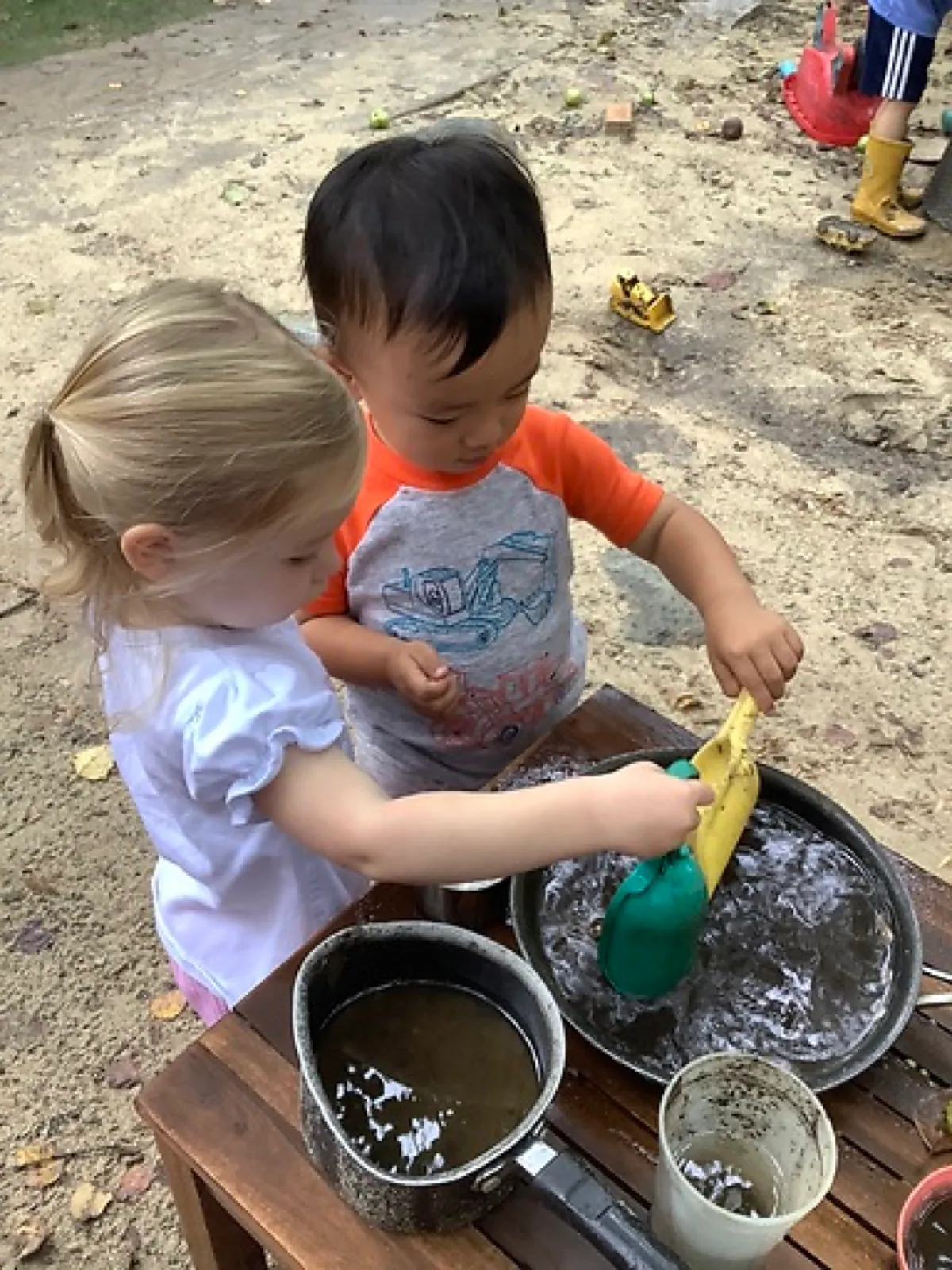 Two toddlers wash sponges in metal pots at LPC's outdoor mud kitchen