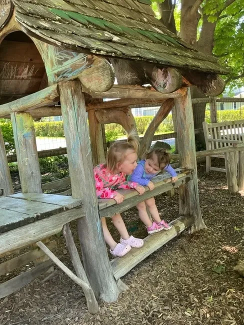 Two toddlers lean out the window of the playground wooden playhouse